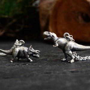 Może przedstawiać: Dwa srebrne zawieszki z dinozaurami na łańcuszkach. Większy zawieszka to Tyrannosaurus Rex, a mniejszy to Triceratops.