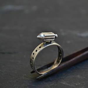 May include: A silver ring with a small, detailed, silver airship charm on top. The ring is held by a pair of tweezers.