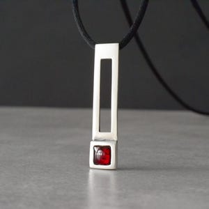 May include: A silver pendant necklace with a rectangular design and a red square gemstone at the bottom. The pendant hangs from a black cord. The necklace is displayed against a gray surface and a dark background.