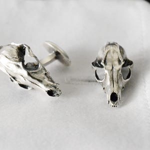 May include: A pair of silver cufflinks shaped like animal skulls. The cufflinks are detailed and realistic.