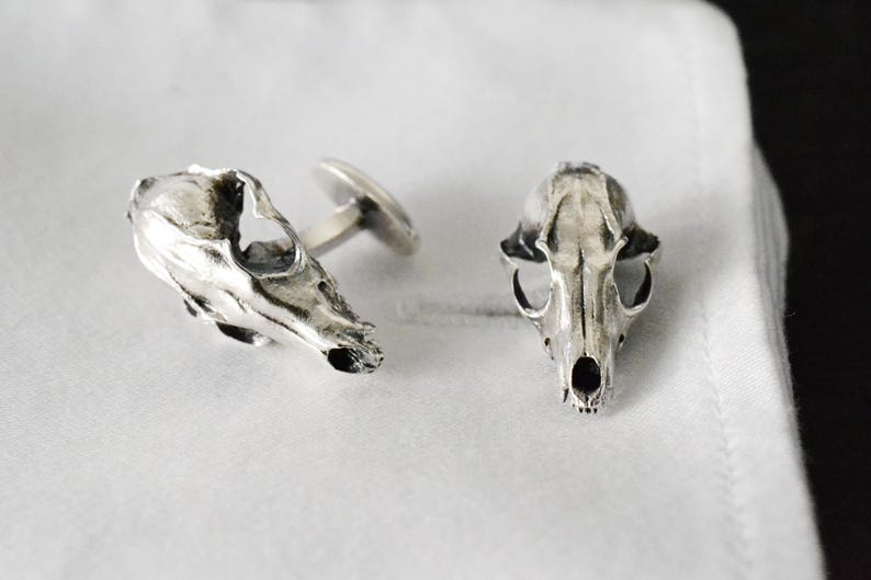 May include: A pair of silver cufflinks shaped like animal skulls. The cufflinks are detailed and realistic.