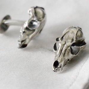 May include: A pair of silver cufflinks shaped like animal skulls. The cufflinks are detailed and realistic.