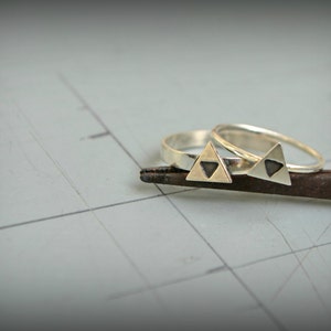 May include: Two silver rings with a black triangle design. The rings are on a rusty metal surface.
