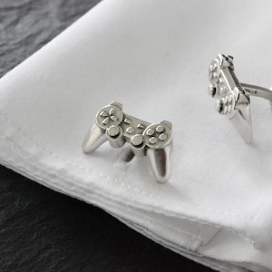 May include: A pair of silver cufflinks shaped like video game controllers. The cufflinks are on a white shirt cuff.