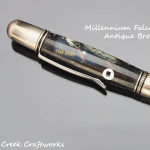 May include: An antique bronze twist pen with a Star Wars theme. The pen features a design with the Millennium Falcon and the Star Wars logo. The pen has a silver clip and accents, and the text "Millennium Falcon Authentic Relic Antique Bronze Twist Pen" is visible.