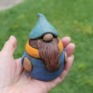 May include: A ceramic gnome figurine with a blue hat dotted with blue circles, a brown beard, and an orange scarf. The gnome wears a blue outfit with brown accents. The figurine is held in a hand against a blurred green background.