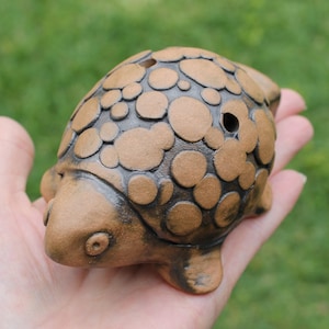 May include: A handcrafted ceramic turtle figurine in shades of brown and black. The turtle has a textured shell with raised circular patterns and small holes. The figurine is held in a hand against a blurred green background.