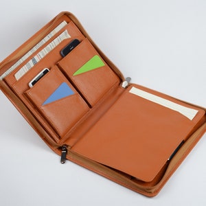 K&ouml;nnte beinhalten: Offenes braunes Leder-Zipper-Portfolio mit mehreren Taschen und einem Notizblock. Die Taschen haben blaue und gr&uuml;ne Akzente.