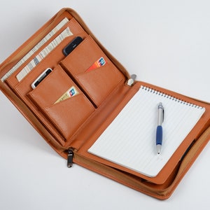 K&ouml;nnte beinhalten: Ein braunes Leder-Zipper-Portfolio mit einem Notizblock, einem Stift und mehreren Taschen f&uuml;r Karten und ein Telefon.