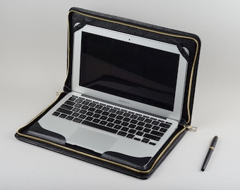 May include: A black leather laptop case with a gold zipper. The case is open and a silver laptop is inside. A black pen is lying on the white surface.