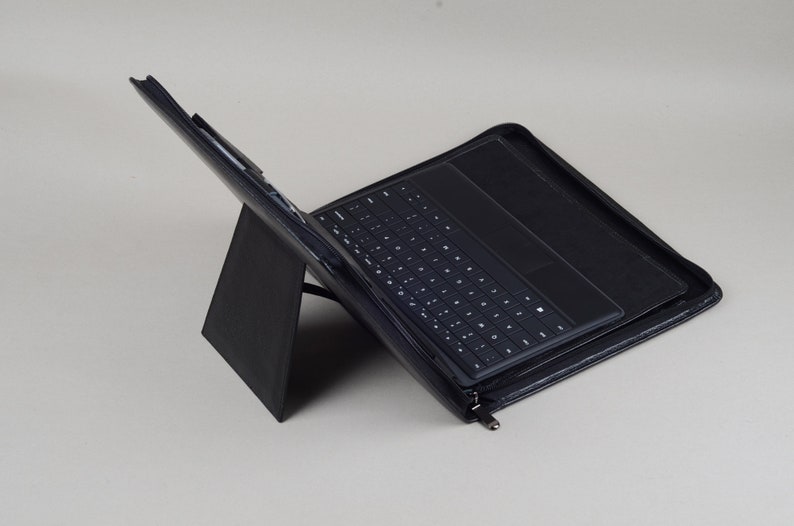 May include: A black leather case with a built-in keyboard and trackpad for a tablet computer. The case is folded to create a stand for the tablet.