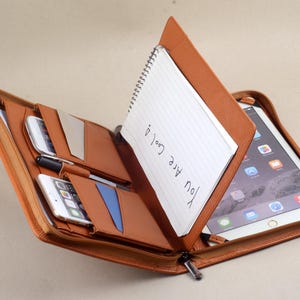 May include: A brown leather portfolio with a zipper closure, containing a notepad, a tablet, and two phones. The notepad has the words "You are cool" written on it.