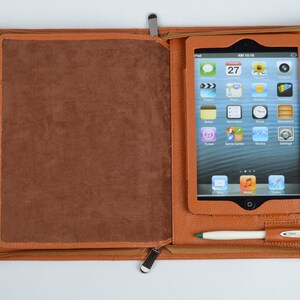 K&ouml;nnte beinhalten: Eine braune Leder-Zipper-Aktentasche mit einem Tablet-Computer im Inneren. Der Tablet-Bildschirm zeigt eine Vielzahl von Apps, darunter eine Uhr, einen Kalender und einen Musikplayer. Es befinden sich zwei Stifte in der Tasche, ein wei&szlig;er und ein gr&uuml;ner.