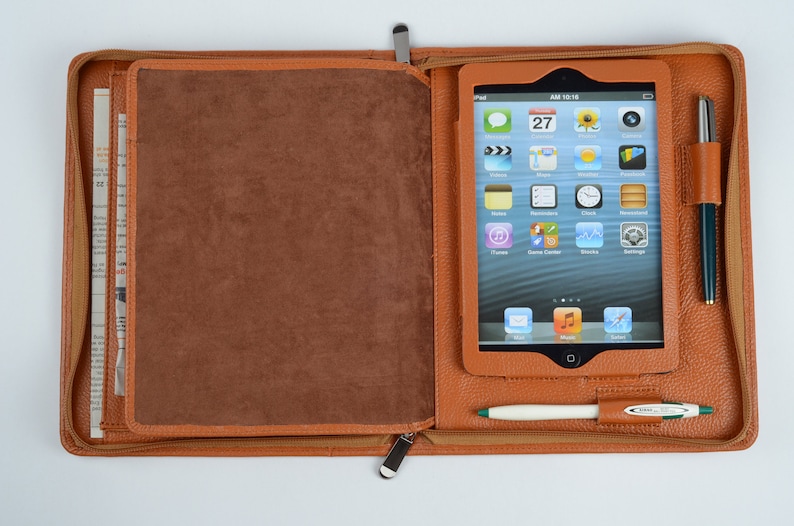 K&ouml;nnte beinhalten: Eine braune Leder-Zipper-Aktentasche mit einem Tablet-Computer im Inneren. Der Tablet-Bildschirm zeigt eine Vielzahl von Apps, darunter eine Uhr, einen Kalender und einen Musikplayer. Es befinden sich zwei Stifte in der Tasche, ein wei&szlig;er und ein gr&uuml;ner.