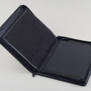 Puede incluir: Una funda de cuero negro con cierre de cremallera, diseñada para contener una tableta. La funda está abierta, revelando la pantalla de la tableta.