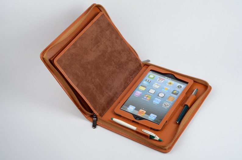 K&ouml;nnte beinhalten: Eine braune Ledertasche mit Rei&szlig;verschluss, in der sich ein Tablet und ein Stift befinden. Das Tablet zeigt einen Startbildschirm mit verschiedenen Apps an.