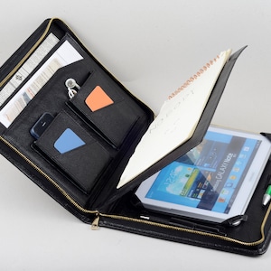 以下が含まれることがあります： 金色のジッパーが付いた黒い革製ポートフォリオケース。タブレット、メモ帳、ポケットが現れるように開いています。タブレットは青と白の画面を表示します。ポートフォリオには、ペン、カード、スマートフォンも収納されています。