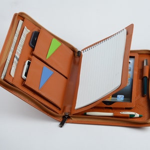 K&ouml;nnte beinhalten: Ein braunes Leder-Zipper-Portfolio mit einem Notizblock, einem Stift und einem Tablet im Inneren. Das Portfolio hat mehrere Taschen und F&auml;cher f&uuml;r die Organisation.