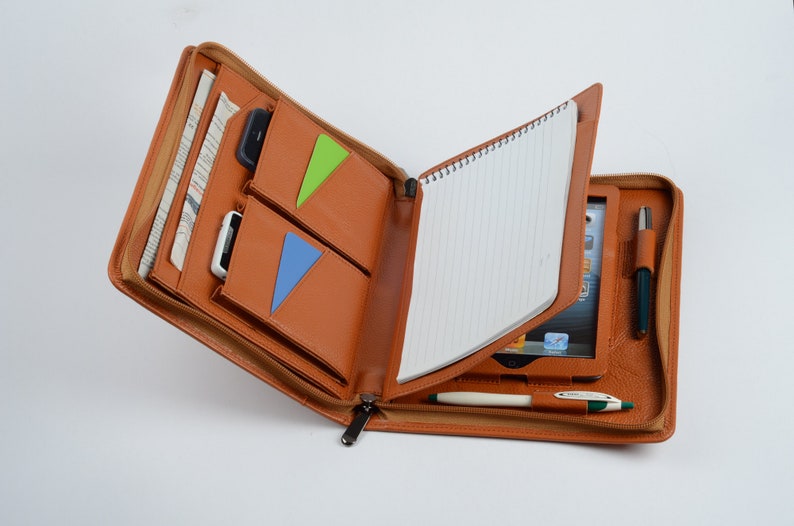 K&ouml;nnte beinhalten: Ein braunes Leder-Zipper-Portfolio mit einem Notizblock, einem Stift und einem Tablet im Inneren. Das Portfolio hat mehrere Taschen und F&auml;cher f&uuml;r die Organisation.