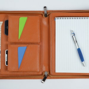 K&ouml;nnte beinhalten: Ein braunes Leder-Zipper-Portfolio mit mehreren Taschen, einem Notizblock und einem Stift. Das Portfolio hat einen Rei&szlig;verschluss und einen Metall-Rei&szlig;verschluss.