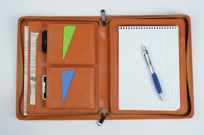 K&ouml;nnte beinhalten: Ein braunes Leder-Zipper-Portfolio mit mehreren Taschen, einem Notizblock und einem Stift. Das Portfolio hat einen Rei&szlig;verschluss und einen Metall-Rei&szlig;verschluss.