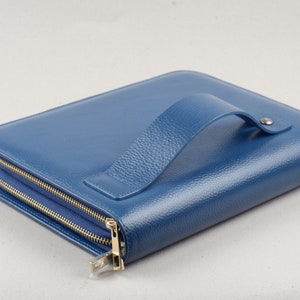 May include: A blue leather zippered portfolio with a handle. The portfolio is closed and lying on a white surface.