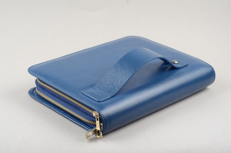 May include: A blue leather zippered portfolio with a handle. The portfolio is closed and lying on a white surface.