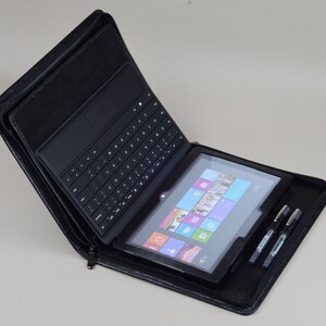 May include: A black leather case with a zipper closure, holding a black tablet with a keyboard attached and a stylus pen. The tablet screen shows the Windows Start menu.