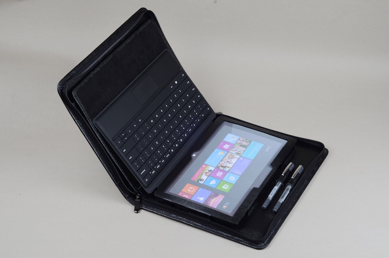 May include: A black leather case with a zipper closure, holding a black tablet with a keyboard attached and a stylus pen. The tablet screen shows the Windows Start menu.