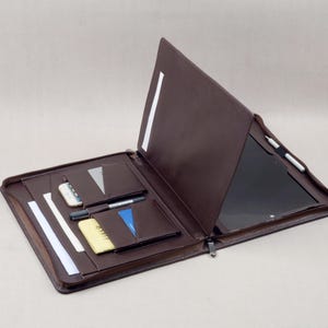 Puede incluir: Una carpeta portafolio de cuero marrón oscuro con soporte integrado para tableta, múltiples bolsillos interiores para tarjetas y documentos, un lazo para bolígrafo y cierre de cremallera. El interior contiene un smartphone, una libreta y un lápiz óptico.