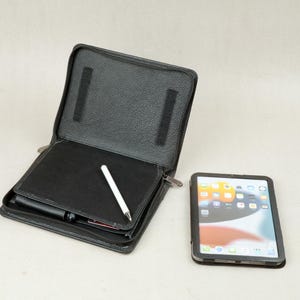 Puede incluir: Funda de tableta de cuero negro con cremallera, abierta para revelar un interior negro, un lápiz óptico blanco y una tableta. La tableta está en una funda negra. La funda tiene dos correas negras en el interior.