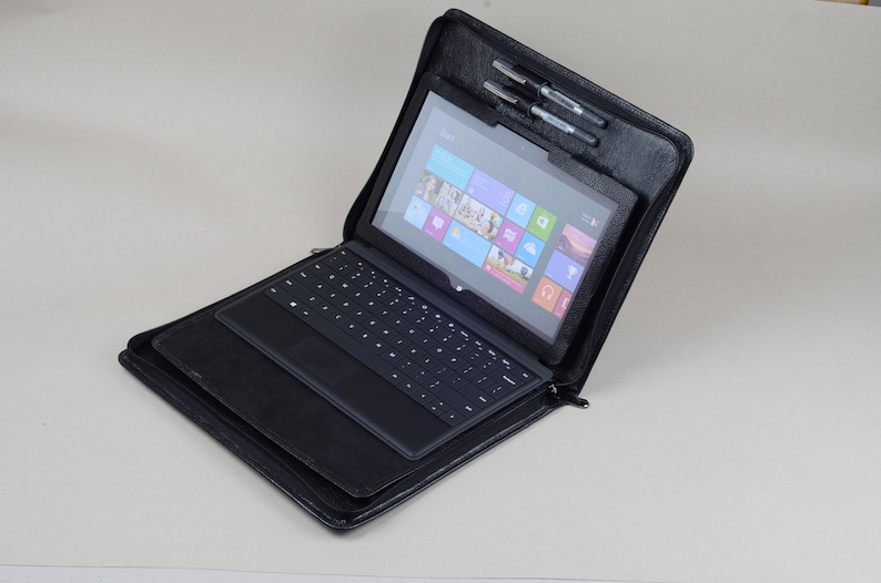 May include: A black leather laptop case with a zipper closure. The case is open and a black laptop with a keyboard and a touchscreen is inside. The laptop screen is on and shows the Windows desktop.
