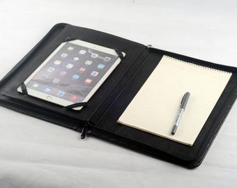 May include: Black leather portfolio with a zipper closure. The portfolio has a built-in tablet holder and a notepad with lined paper. A pen is lying on the notepad.
