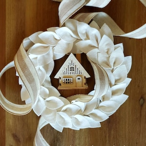 May include: A white felt leaf wreath with a small wooden house in the center. The wreath is on a wooden surface with a tan and white striped ribbon. The text "listing is for wreath only decorative props not included" is at the bottom of the image.