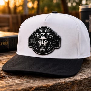 May include: A white baseball cap with a black brim featuring a central black patch. The patch displays a graphic illustration of a face wearing a crown of thorns with the text JOHN 3:16 printed on either side of the central image.