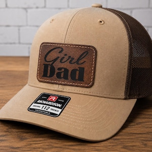 May include: A tan and brown trucker hat with a mesh back. The hat features a leather patch with the words "Girl Dad" in black lettering. The hat has a curved brim and an adjustable strap. The brand label "Richardson 112 Trucker" is visible.