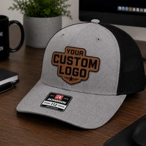 May include: A grey and black trucker cap with a custom logo patch. The patch is brown and features the words "YOUR CUSTOM LOGO" and a star design. The cap has a curved peak and a mesh back. It rests on a wooden surface.