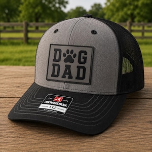 May include: A gray and black trucker hat with the words "DOG DAD" in a square patch. The hat has a black brim and mesh back. The Richardson brand label is visible.