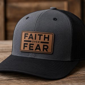 May include: A charcoal gray and black trucker hat featuring a rectangular brown leather patch on the front panel. The patch is embossed with the black text FAITH OVER FEAR in a bold, sans-serif font. The hat has a solid front and mesh back.