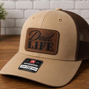 May include: A tan and brown trucker hat featuring a rectangular brown leather patch on the front with the text Dad LIFE embossed in black. The hat has a mesh back, a curved brim, and a black sticker on the brim that reads RICHARDSON 112 TRUCKER.