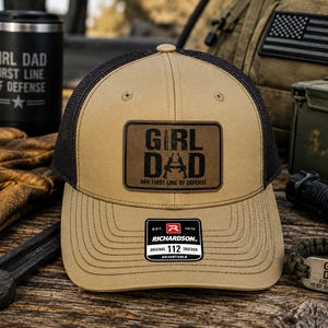 May include: A tan and black trucker hat featuring a rectangular brown patch on the front with the text GIRL DAD and the phrase HER FIRST LINE OF DEFENSE. The hat includes a black mesh back and a Richardson 112 sticker on the curved brim.