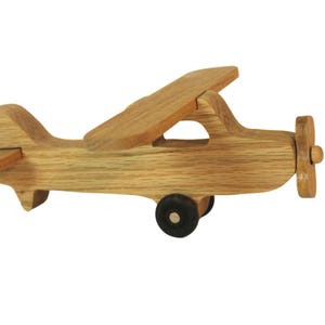 May include: A wooden toy airplane with a propeller, wings, and tail. The toy is made of light-colored wood and has a single black wheel. The propeller is at the front of the plane.