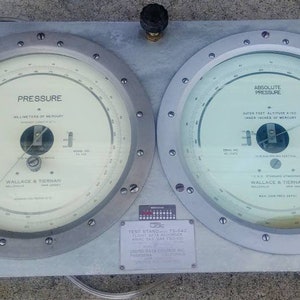 May include: A silver metal instrument panel with two circular gauges. The left gauge reads "PRESSURE" and the right gauge reads "ABSOLUTE PRESSURE". Both gauges have a white face with black markings and a needle. The panel has a black knob on top.