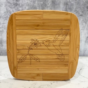 May include: A natural bamboo cutting board with a laser-etched design of a hummingbird feeding from a flower.