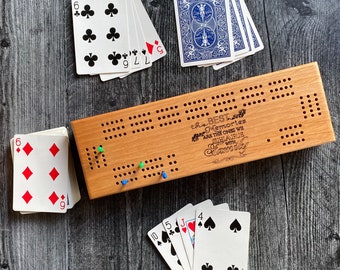 May include: A wooden cribbage board with a light brown stain. The board has a white inscription that reads "The best memories are the ones we share with family". There are playing cards scattered around the board, including a deck of blue playing cards and a deck of standard playing cards.