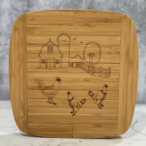 May include: A brown bamboo cutting board with a farmhouse scene engraved on it. The scene includes a barn, silo, fence, tree, and three chickens.