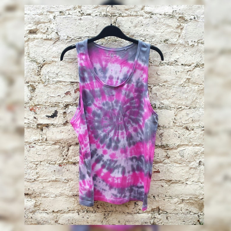 A tie-dye tank top with a pink, gray, and white swirl pattern. The tank top has a scoop neck and is made of a soft, comfortable fabric. The tank top is hanging on a hanger against a brick wall background.
