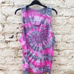 A tie-dye tank top with a pink and gray swirl design. The tank top has a scoop neck and is made of a soft, comfortable fabric. The tank top is hanging on a black hanger against a white brick wall.