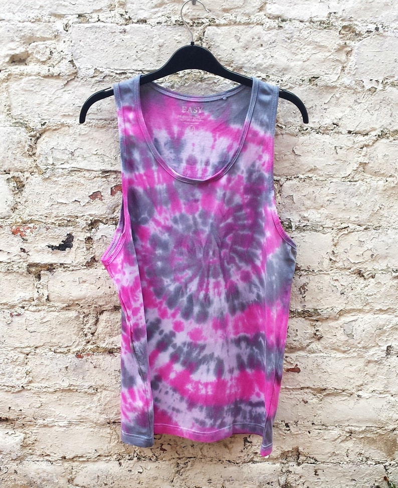 A tie-dye tank top with a pink and gray swirl design. The tank top has a scoop neck and is made of a soft, comfortable fabric. The tank top is hanging on a black hanger against a white brick wall.