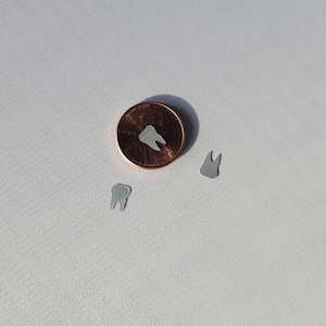 May include: Three small, white tooth-shaped paper cutouts on a white surface. A penny is in the center of the image for scale.